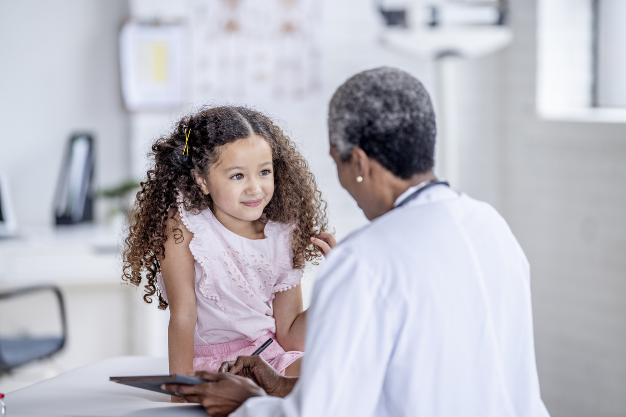 A sweet little girl of Hispanic decent, sits up on an exam table during a routine check-up. She is dressed casually and her doctor is wearing a white lab coat as the appointment progresses and she records information on a tablet.